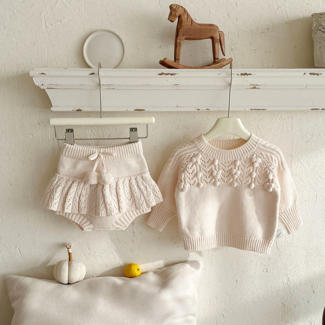 Children's white sweater and shorts set on a white surface with a wooden rocking horse in the background.