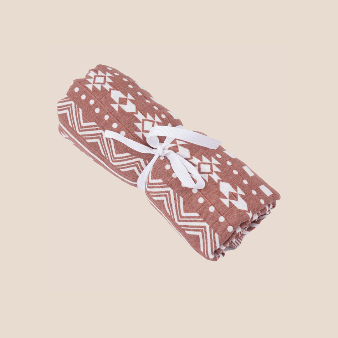 Rolled bamboo muslin swaddle blanket in earthy terracotta tribal design tied with ribbon – sustainable baby gift item perfect for baby shower gift baskets, personalized baby essentials, custom baby registry picks, and thoughtful new mom gifting ideas.