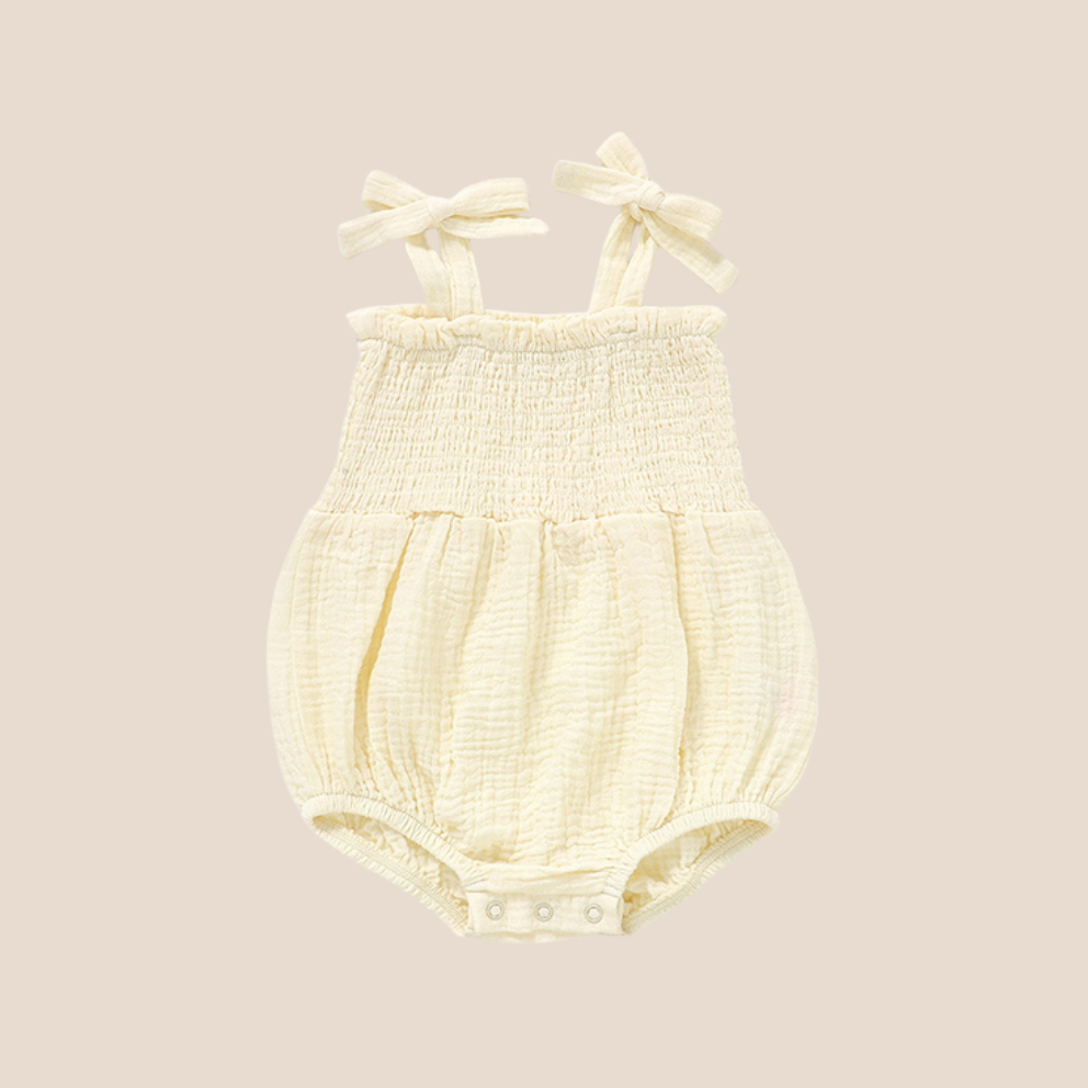 A pale yellow baby romper with a smocked bodice, tie shoulder straps, and snap closures at the crotch, shown against a solid beige background.