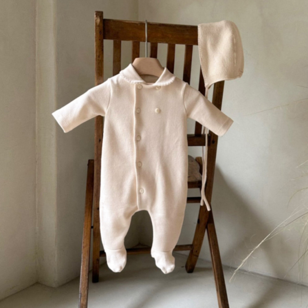 Beige baby onesie with buttons and matching bonnet on a wooden chair against a neutral wall.