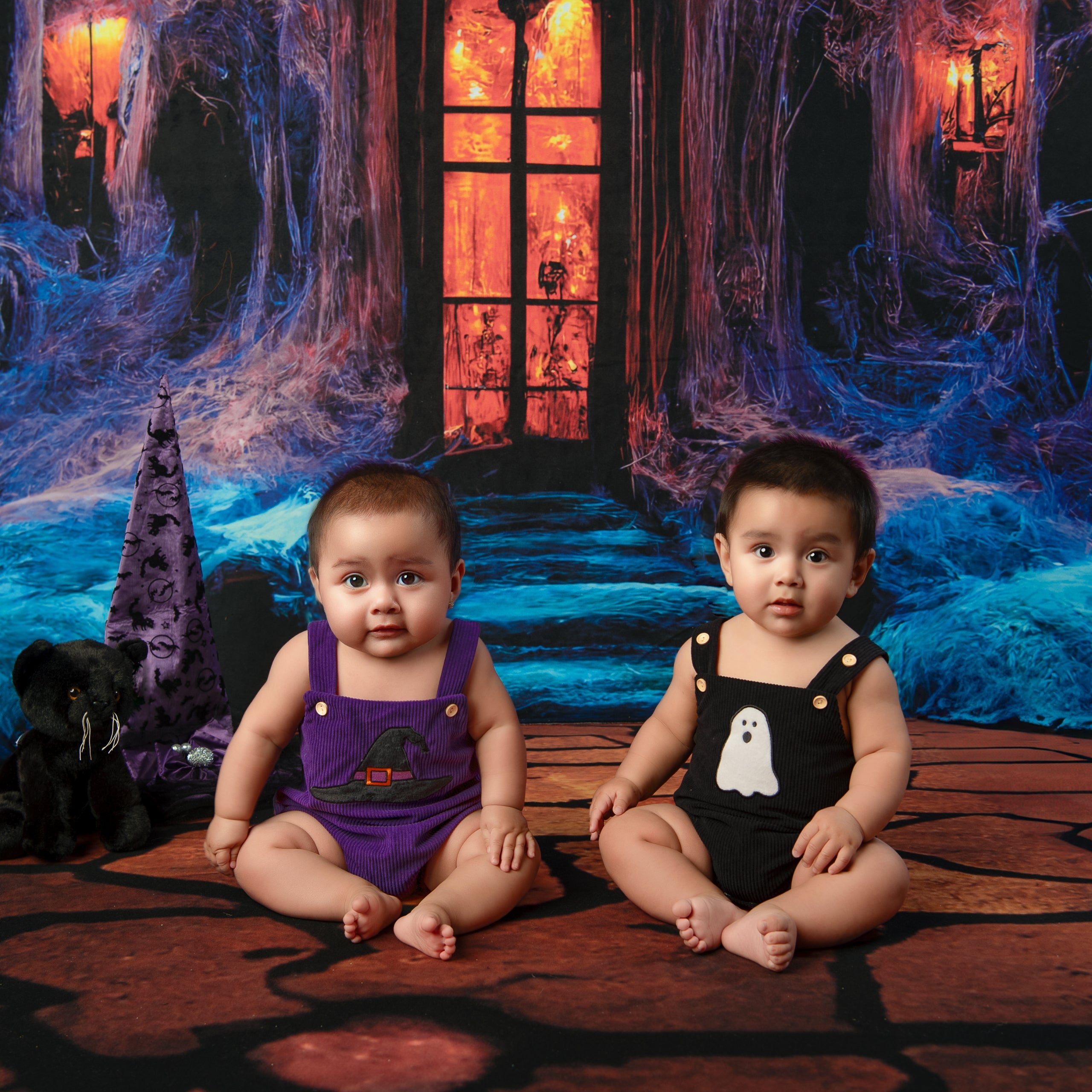 Two children in Halloween-themed outfits sitting on a spider web floor with a spooky backdrop.