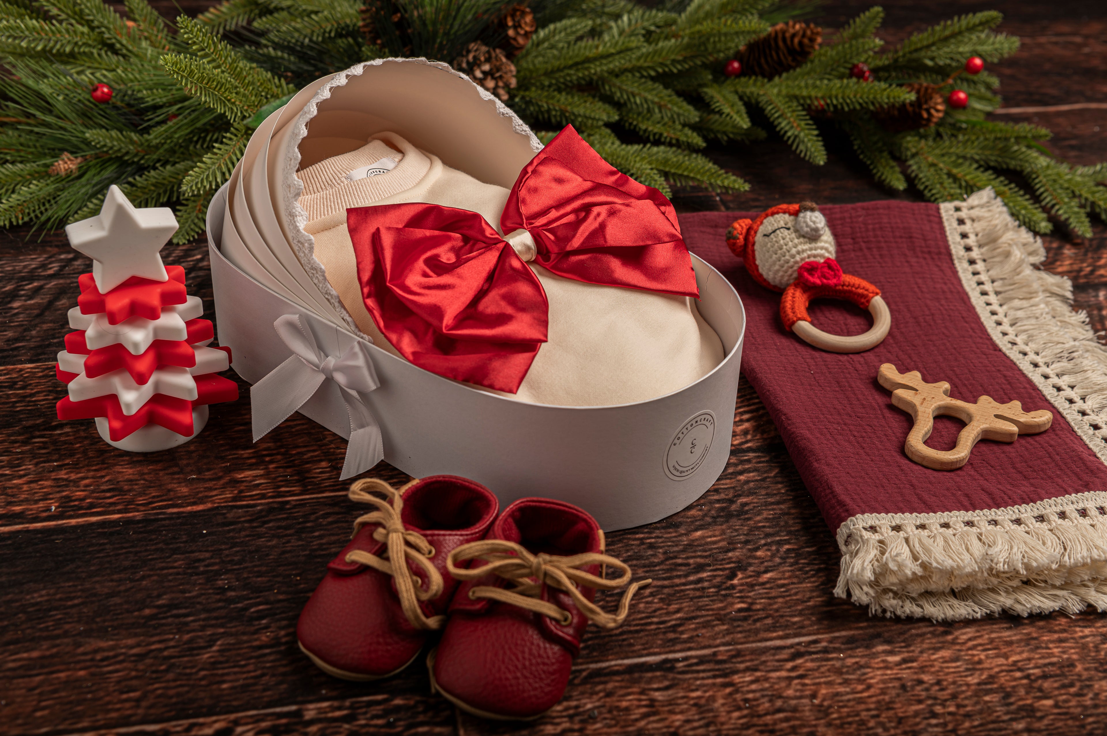 Cotton Craft Luxe Baby Girl Holiday Gift Basket with festive romper, cotton swaddle, leather shoes, hand-knitted rattle, wooden teether, and silicone Christmas tree toy. Red Romper