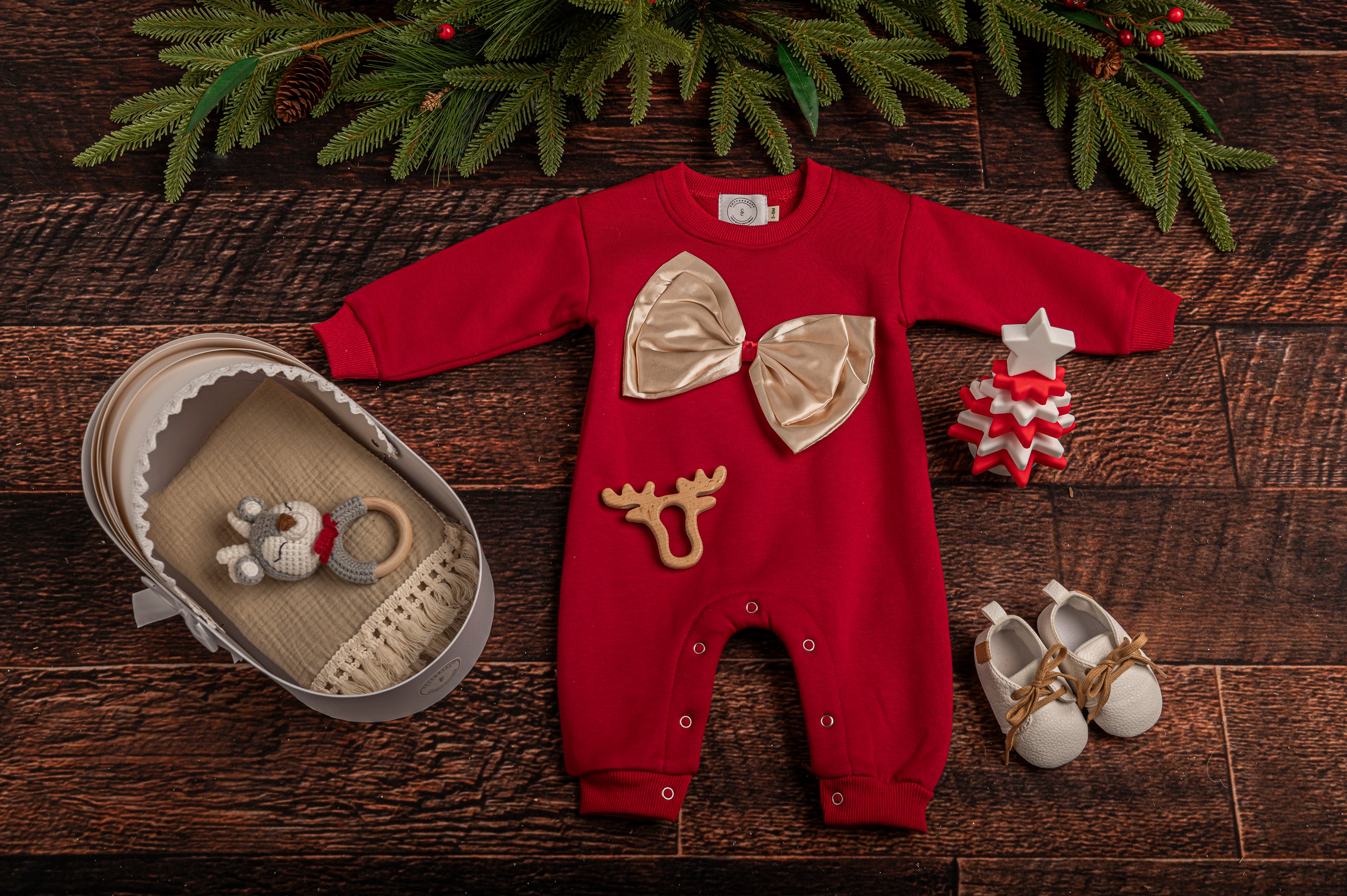 Cotton Craft Luxe Baby Girl Holiday Gift Basket with festive romper, cotton swaddle, leather shoes, hand-knitted rattle, wooden teether, and silicone Christmas tree toy. Flat display