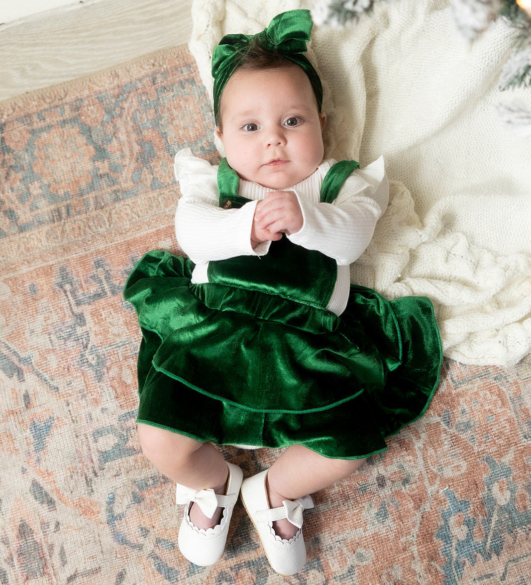Baby in a green and white outfit sitting on a patterned rug with a Christmas tree in the background.
