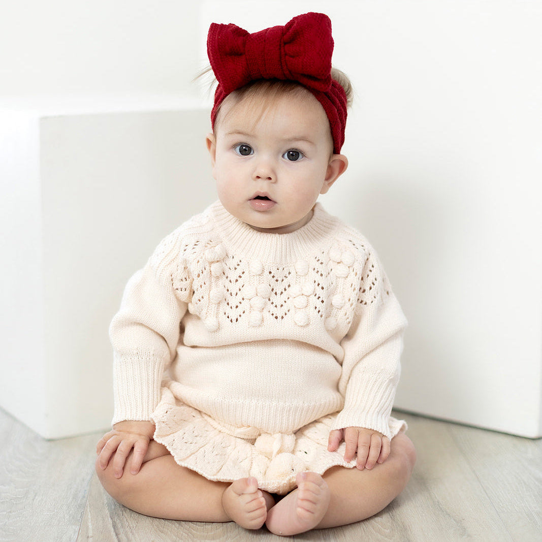 Baby wearing a cream sweater and red headband with a bow, sitting on a white surface.