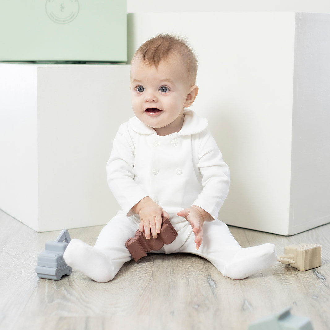 Baby in a white outfit sitting on a light-colored floor with toys around