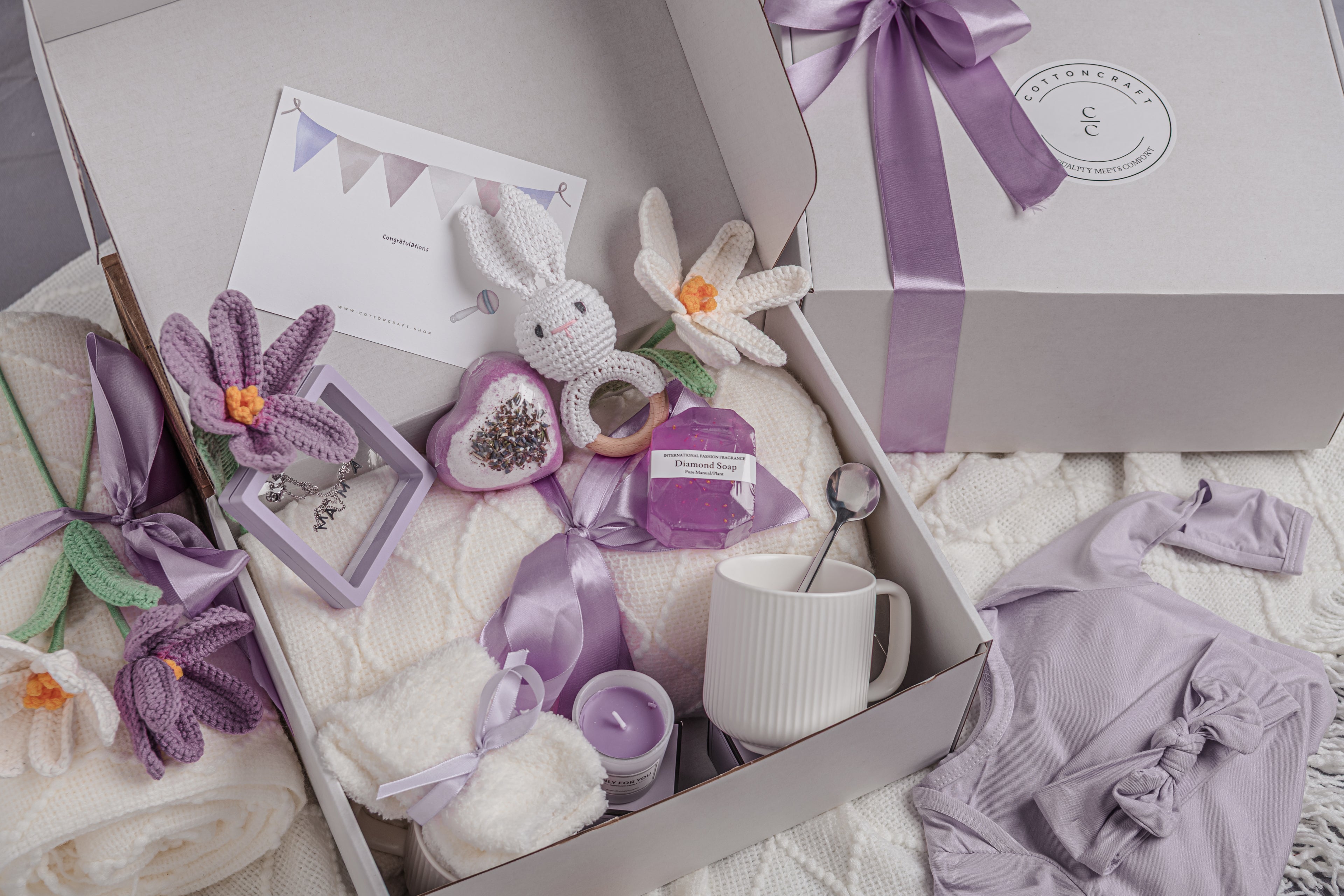 Gift box with baby items including a white rabbit, purple flowers, and a mug, all in a decorative setting.
