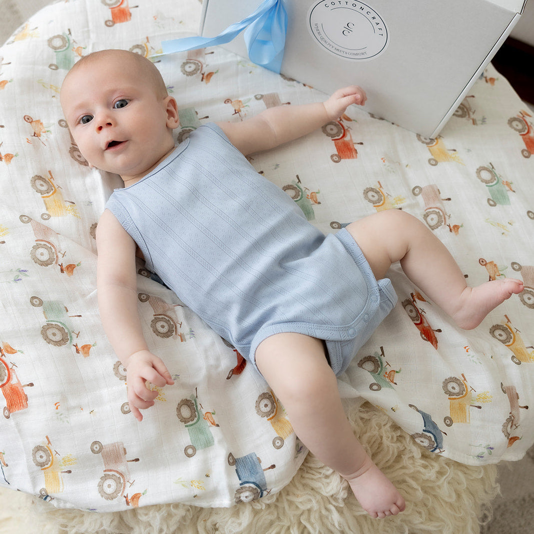 Baby wearing cotton craft romper lying on a truck themed cotton craft organic swaddle blanket with a gift box featuring a blue ribbon in the background from cotton craft baby gifts