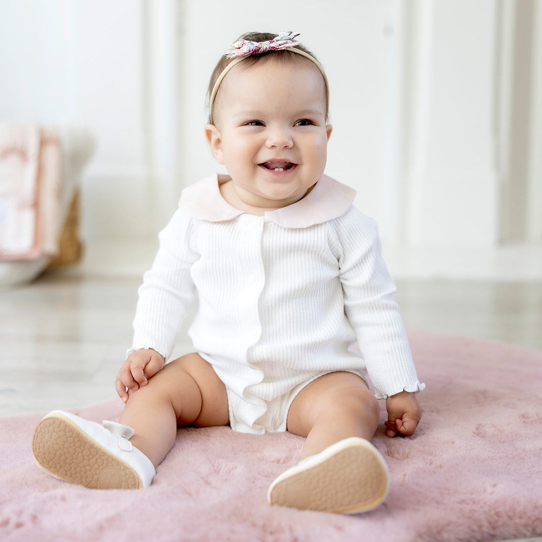 Baby sitting on a pink rug wearing a white outfit with a pink bow.
