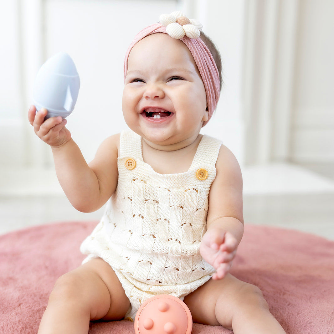 Baby sitting on a pink blanket with toys, wearing a white crocheted romper and pink headband.