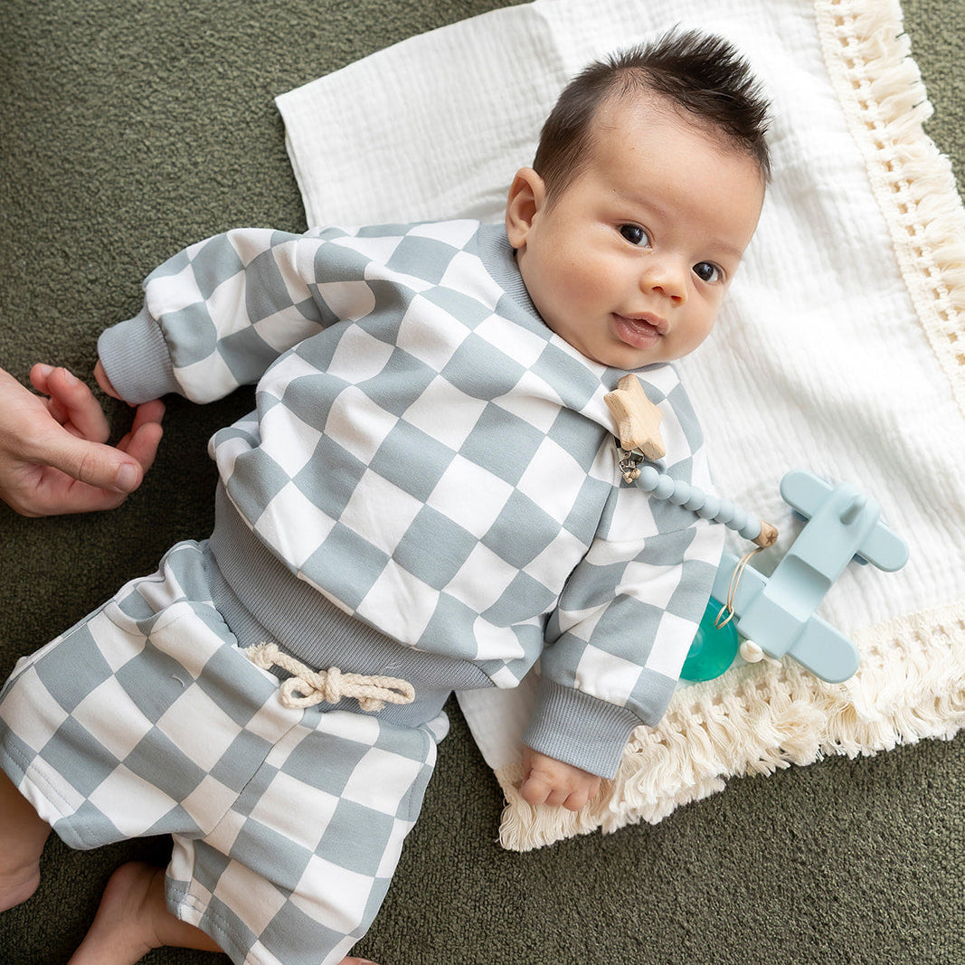 Baby in a checkered outfit lying on a blanket with a toy, surrounded by adult hands.