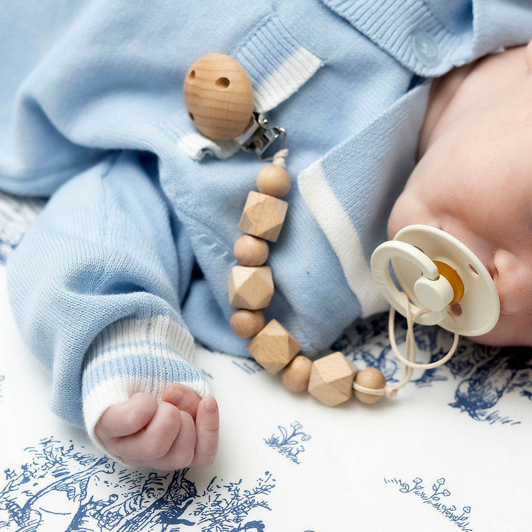 Baby in blue outfit with wooden teething ring and pacifier on floral blanket
