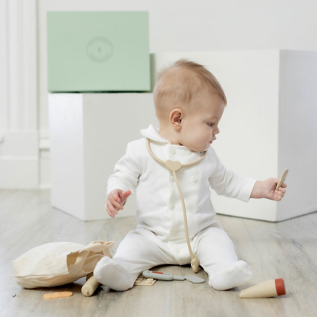 Baby in a white outfit playing with doctor themed silicone toy set on a light wooden floor.