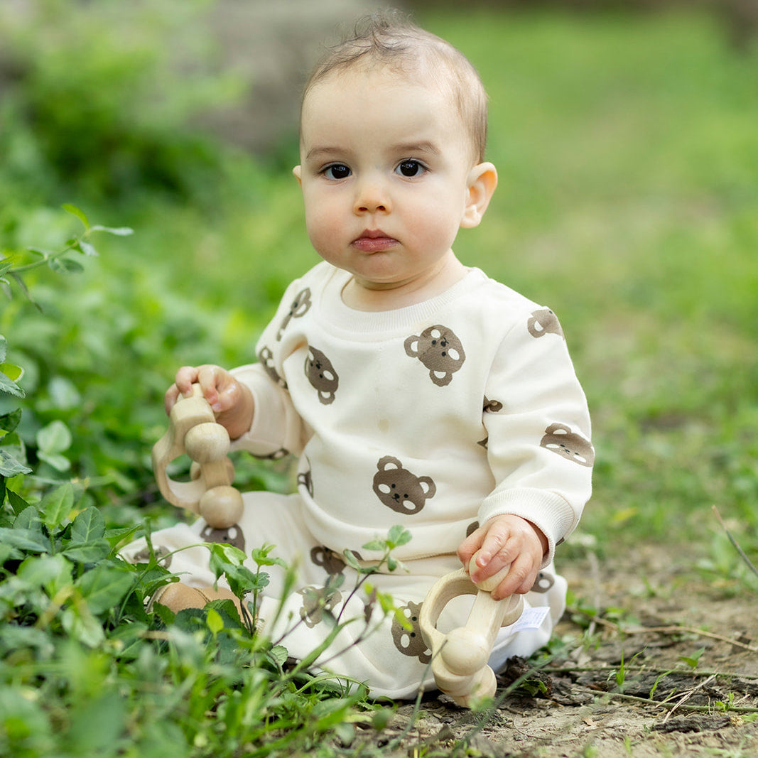 Baby sitting in a grassy area holding wooden toys and wearing cotton craft bear themed cotton 2 piece set