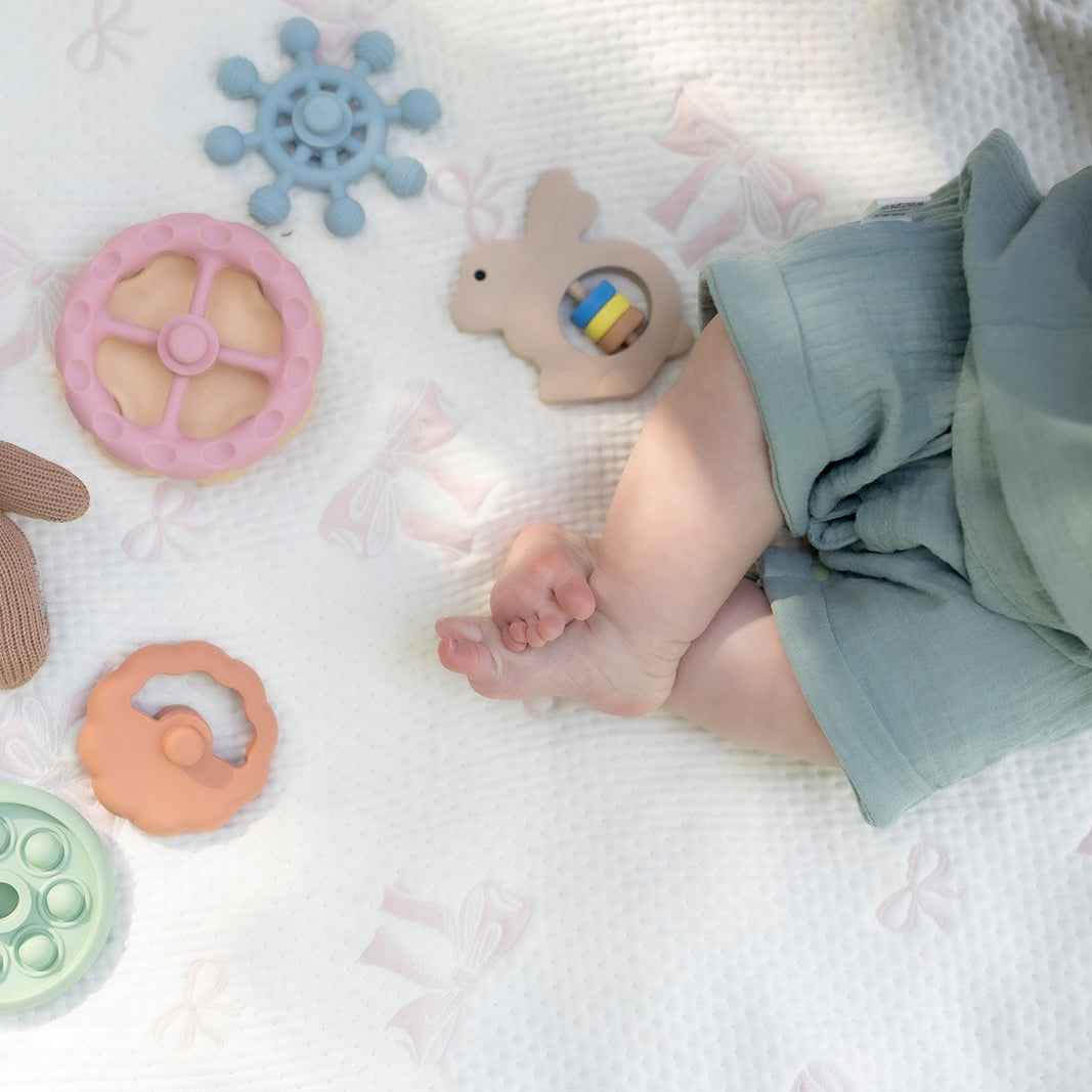 Baby toys including a teddy bear, colorful rings, and a teething ring on a soft surface with a baby's feet visible.