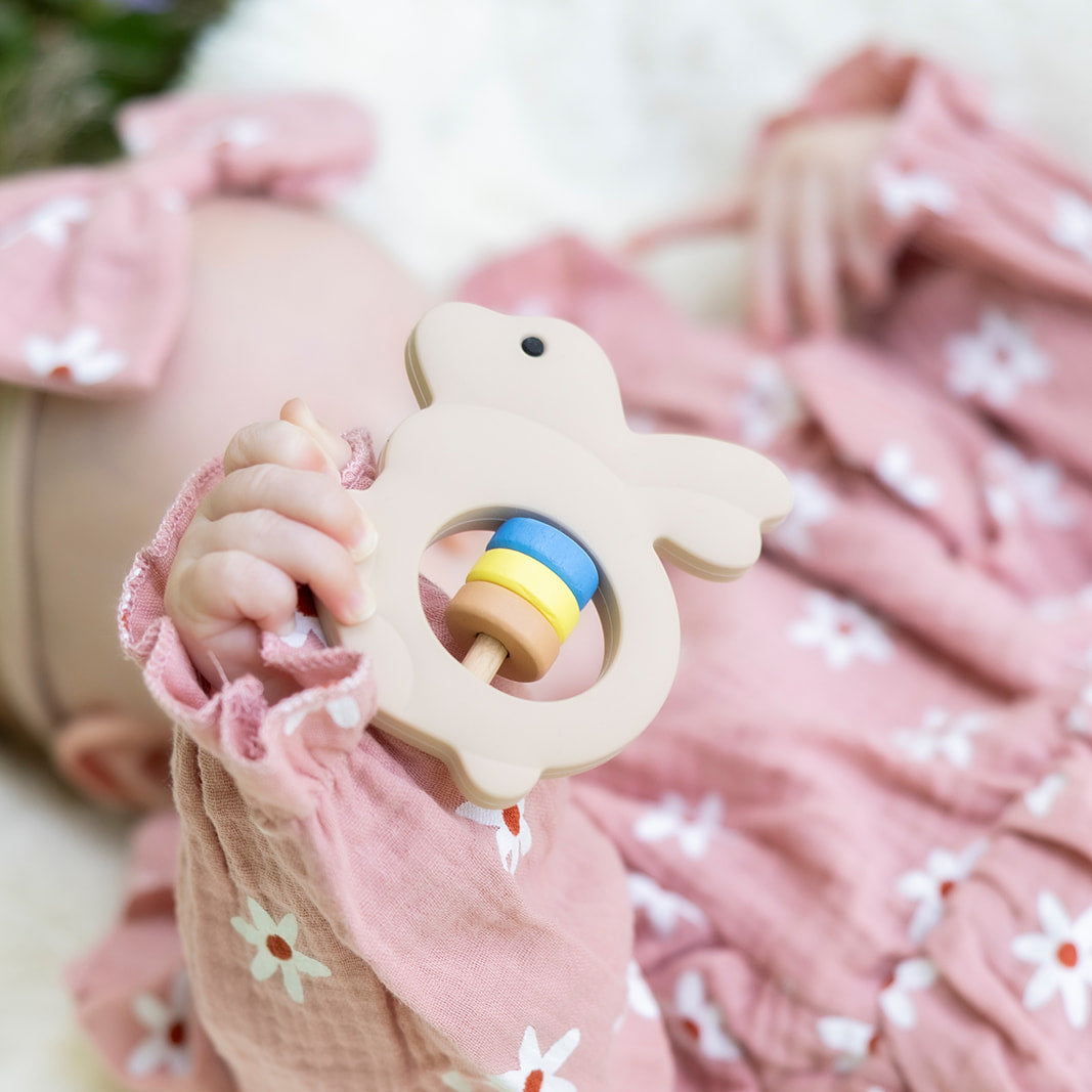 Baby in pink floral outfit holding a teething ring with a rainbow design.