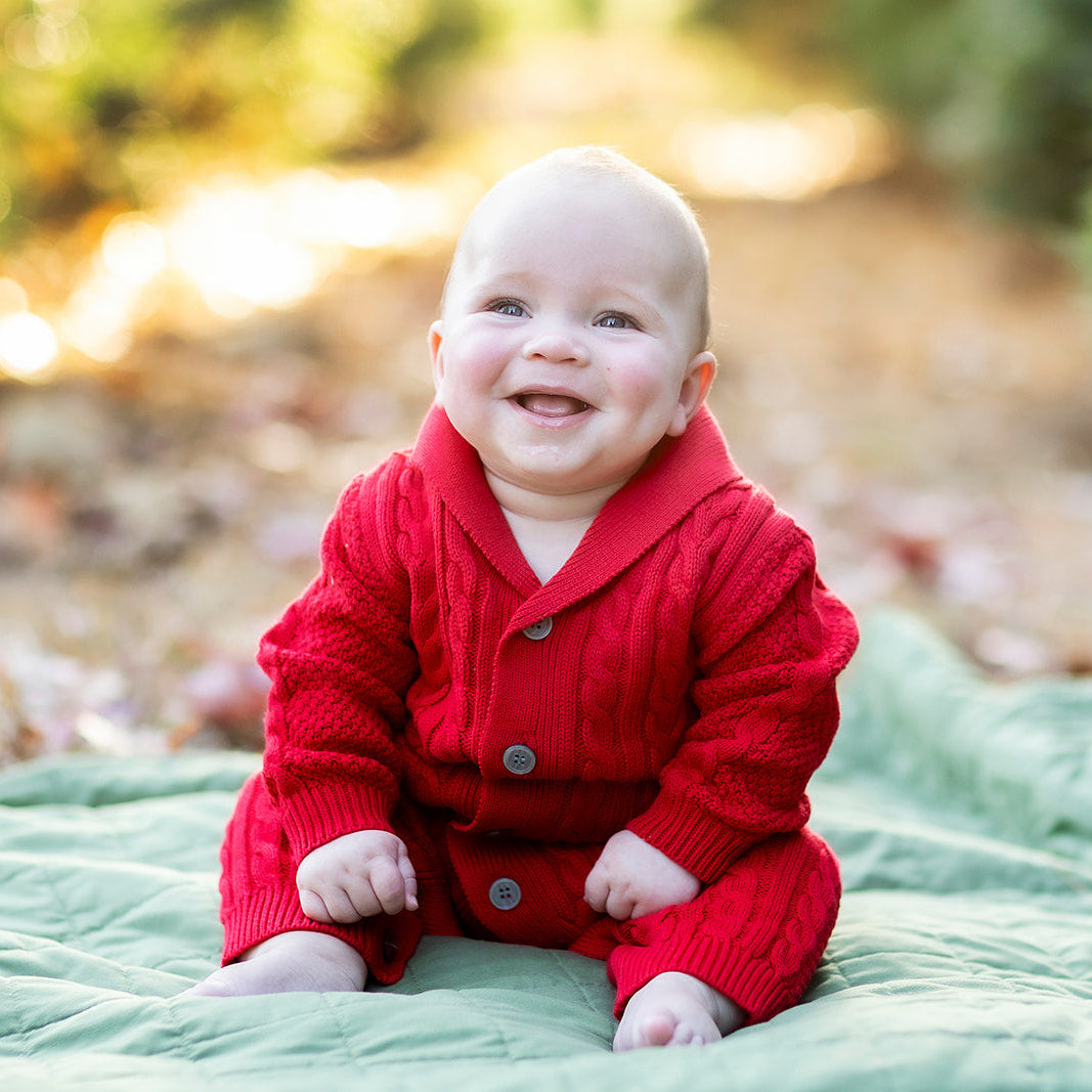 Baby in a red outfit sitting on a blanket outdoors with trees in the background