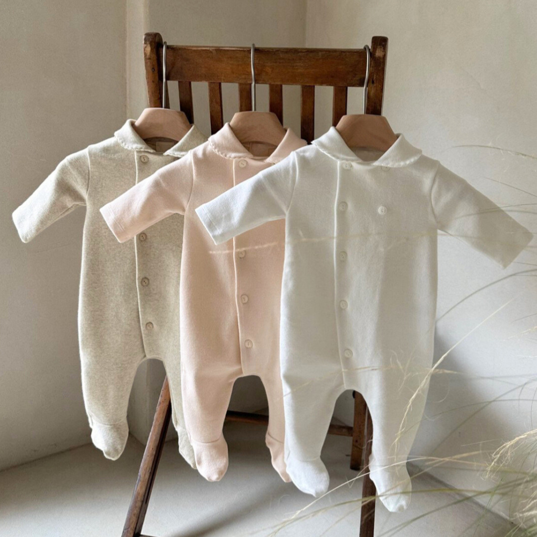 Three baby rompers hanging on a wooden chair against a neutral wall.