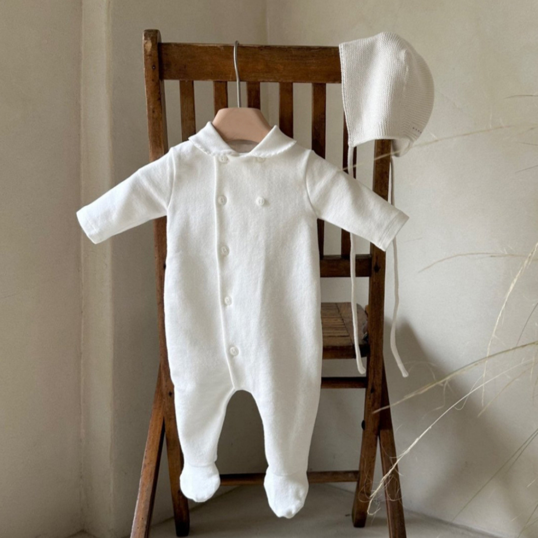 White baby onesie with bonnet on a wooden chair against a neutral wall.