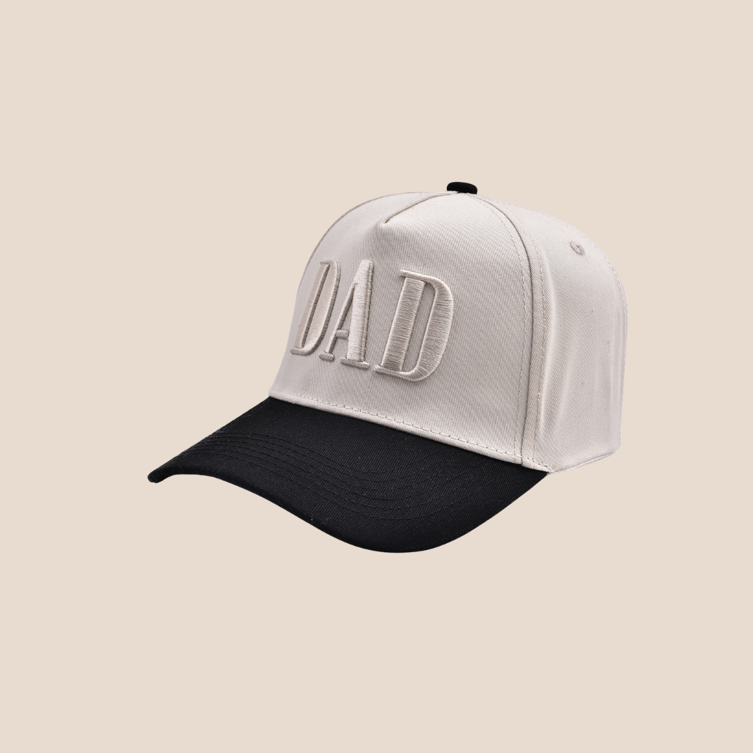 White cap with black bill and 'DAD' embroidery on a beige background