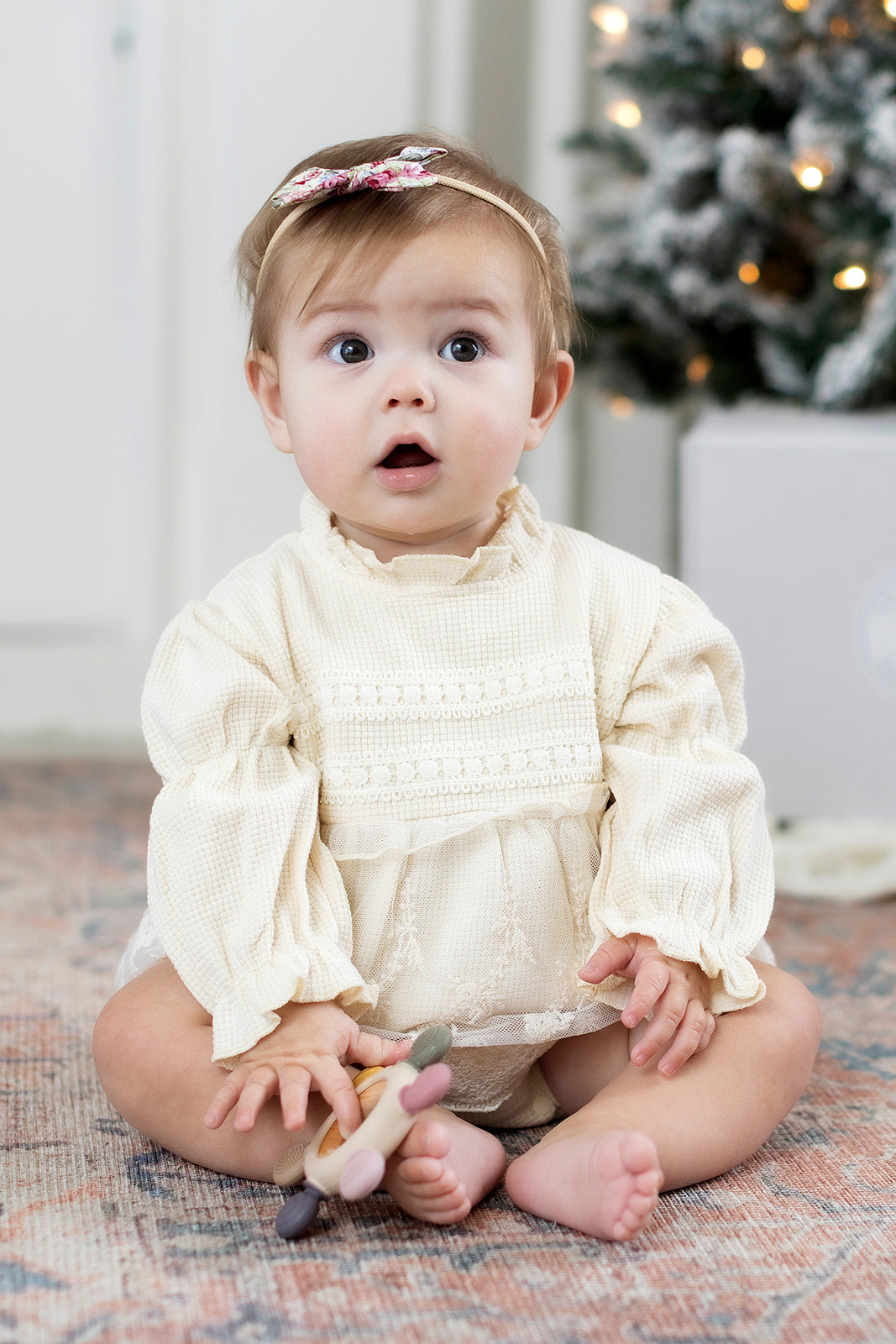 Baby in a white outfit sitting on a patterned rug with a Christmas tree in the background