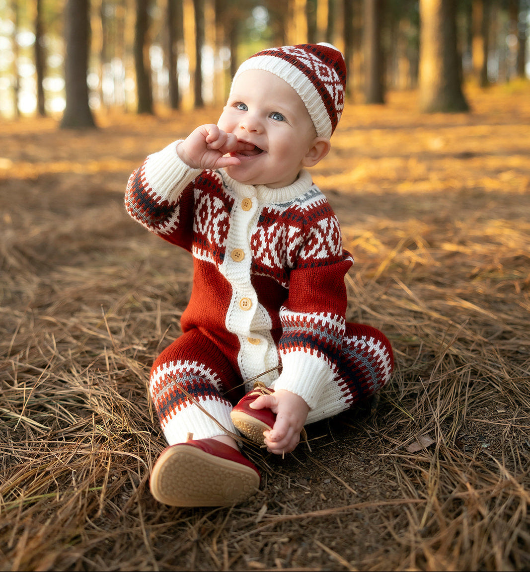 Baby in a red and white patterned outfit sitting on the ground in a forest.