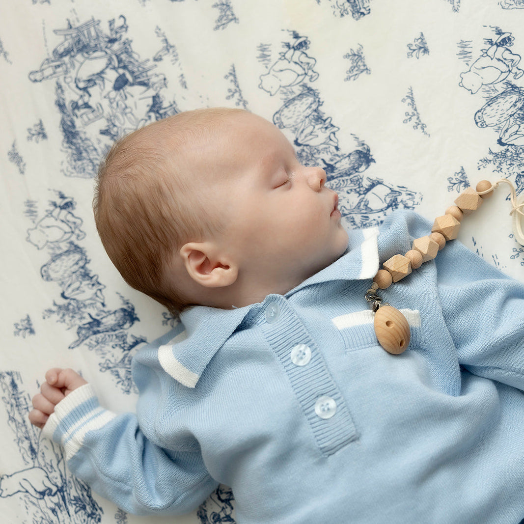 Baby in light blue outfit with a teething toy and pacifier, lying on a patterned surface.