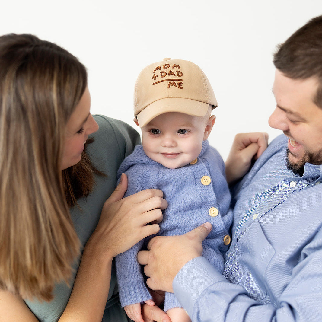 Baby wearing a cap with 'For Dad' text, held by parents against a white background