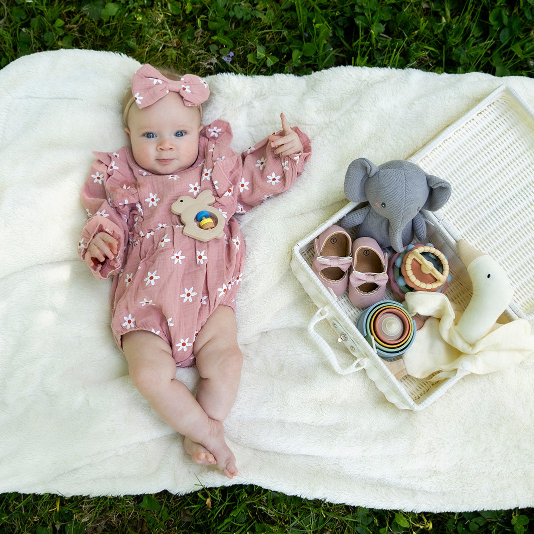 Baby in pink outfit lying on a white blanket with toys and a book