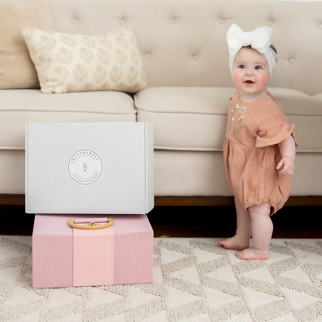 Baby standing next to a pink and gray box on a carpeted floor with a beige couch in the background.